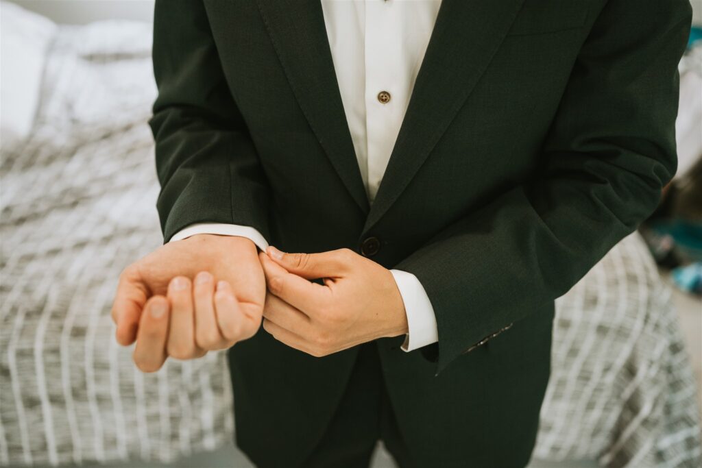 A person in a dark suit adjusts their cufflink, standing in a room with a bed covered by a checkered blanket—preparing for a lakeside elopement.