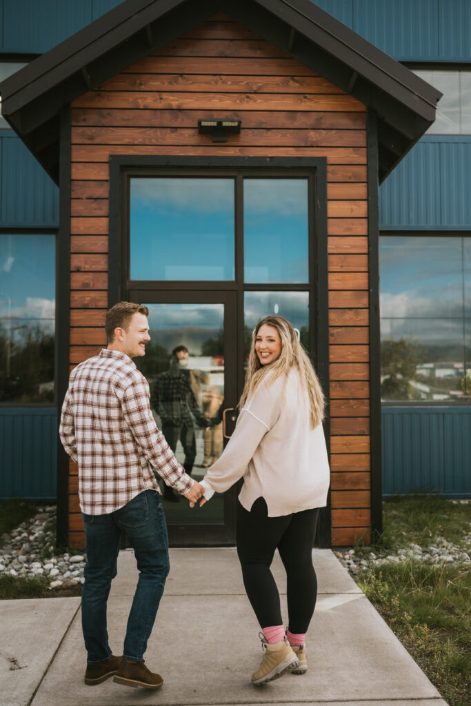 A man and woman holding hands and smiling back while walking toward a building entrance with glass doors and wooden trim, capturing the joy of their lakeside elopement.