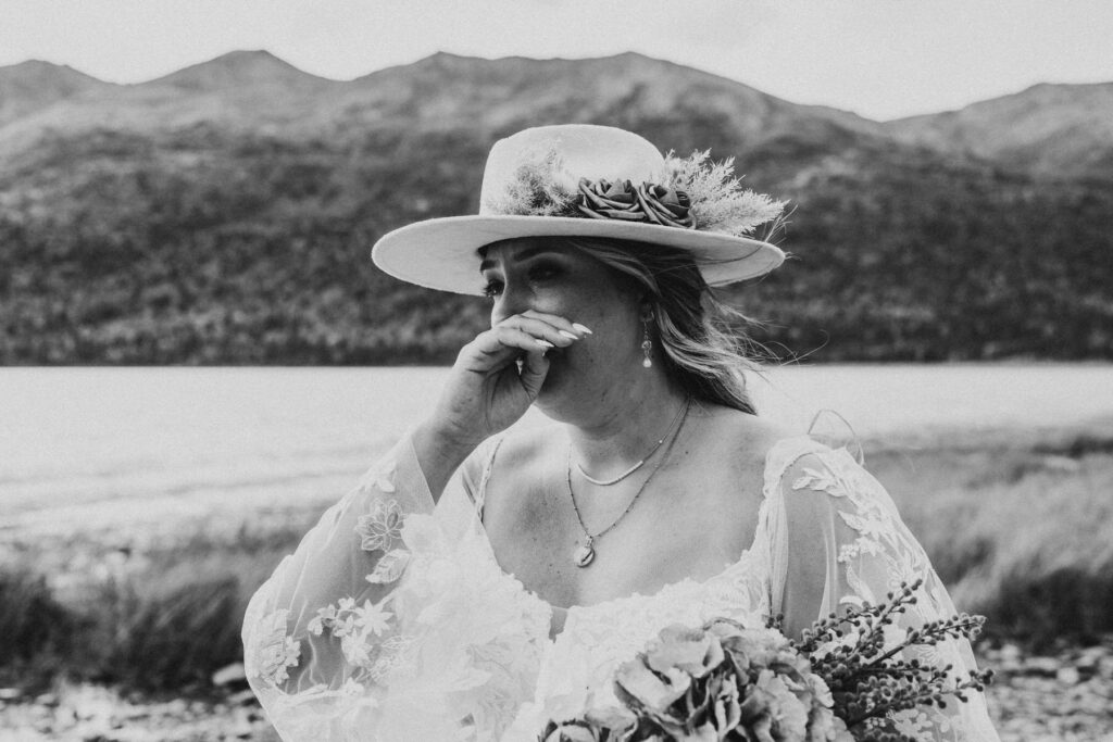 A woman in a white dress and hat stands outdoors by a lake, holding a bouquet and covering her mouth with her hand, capturing the essence of a romantic lakeside elopement with mountains in the background.