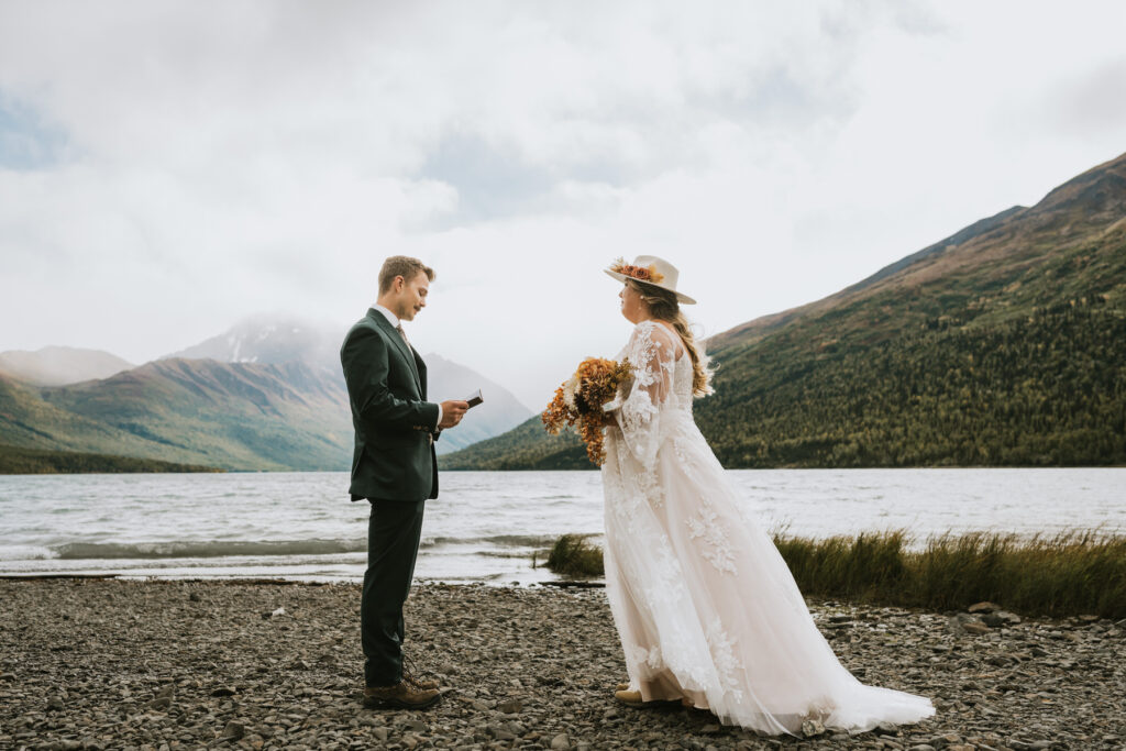 A bride and groom share a heartfelt moment during their lakeside elopement, standing on a rocky shore with mountains behind them; the groom reads from a card while the bride, in a white dress and hat, holds her bouquet.