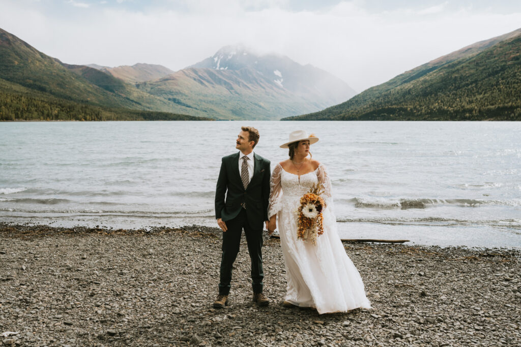 A bride and groom stand on a rocky lakeshore during their lakeside elopement, with mountains and a cloudy sky in the background.