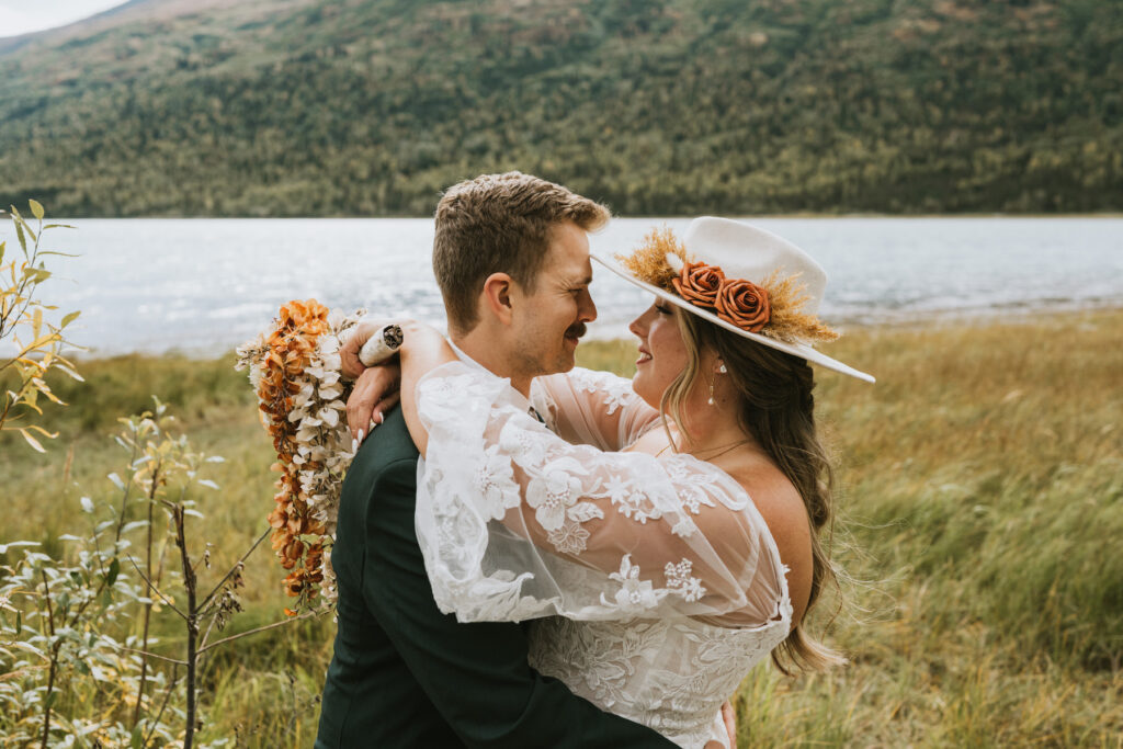 A bride and groom embrace outdoors during their lakeside elopement, with mountains and greenery in the background. The bride wears a white dress and a hat adorned with flowers.