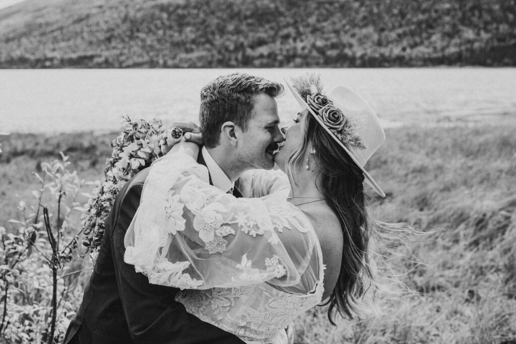 A couple dressed in wedding attire shares a joyful embrace and smile during their lakeside elopement, with hills in the background.