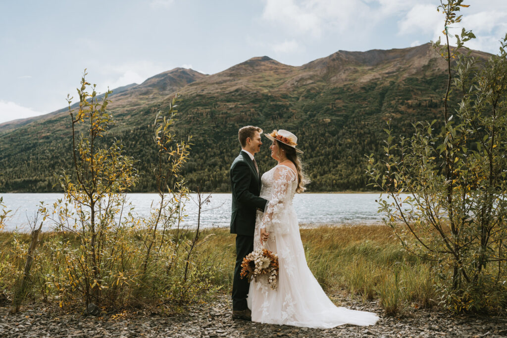 A bride and groom stand facing each other during a lakeside elopement, surrounded by greenery, with mountains in the background under a partly cloudy sky.