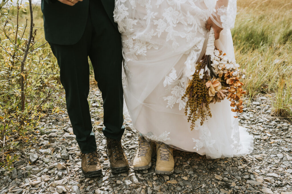 A bride and groom stand on rocky ground by the water; the bride holds a bouquet and both wear hiking boots with formal wedding attire, capturing the spirit of a lakeside elopement.