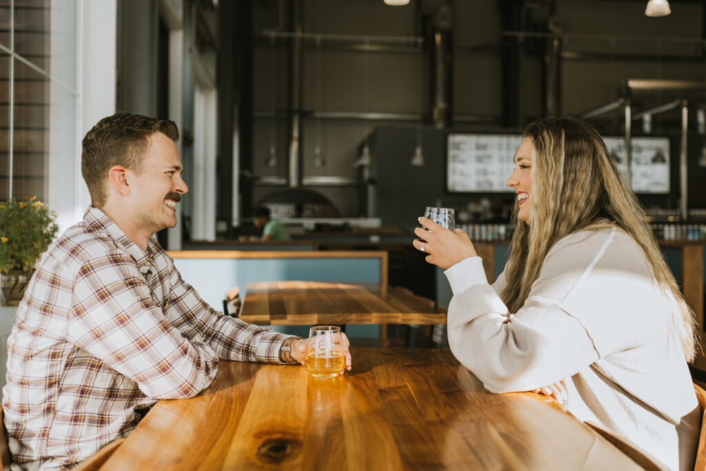 Two people sit across from each other at a wooden table in a bright, modern cafe, smiling and holding drinks as they reminisce about their recent lakeside elopement.