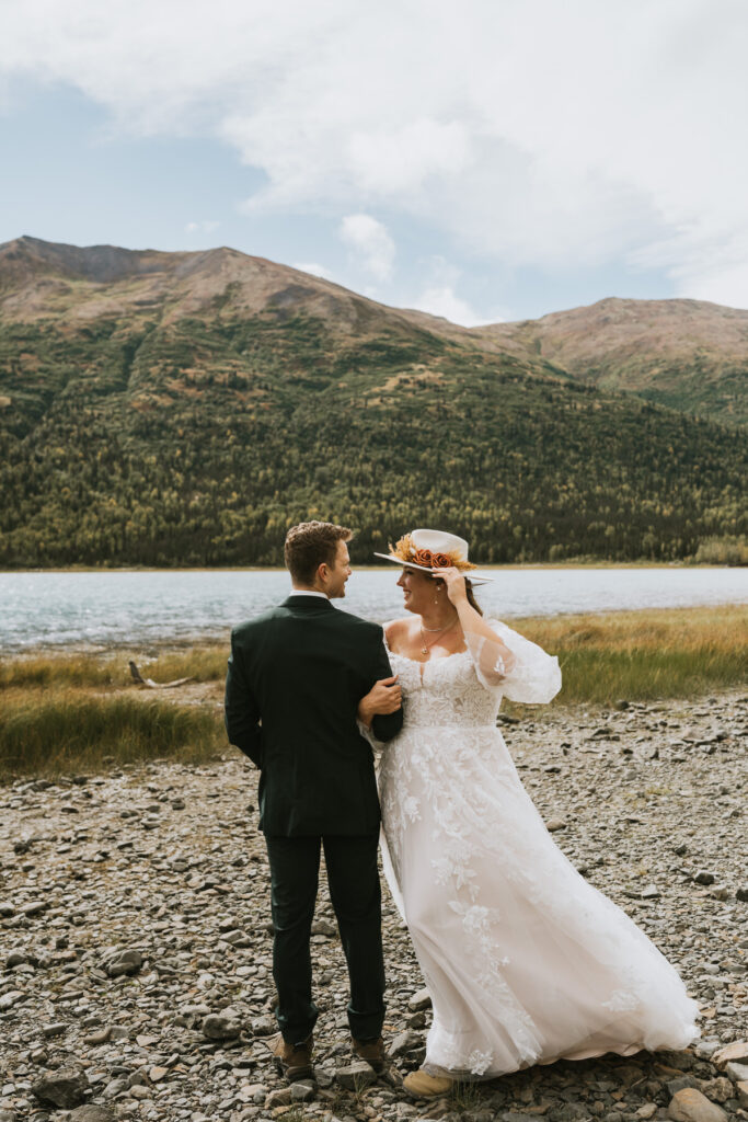 A bride and groom stand together on a rocky lakeshore with mountains and forest in the background, sharing a quiet moment during their lakeside elopement as they look lovingly at each other.