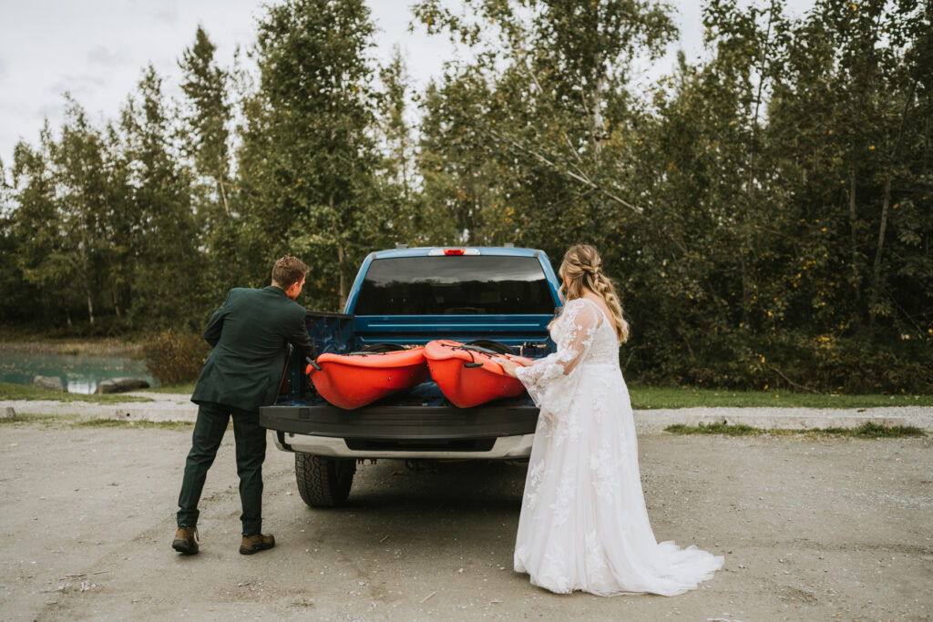 A man in a suit and a woman in a wedding dress load two red kayaks into the back of a blue pickup truck, preparing for their lakeside elopement in a wooded outdoor area.