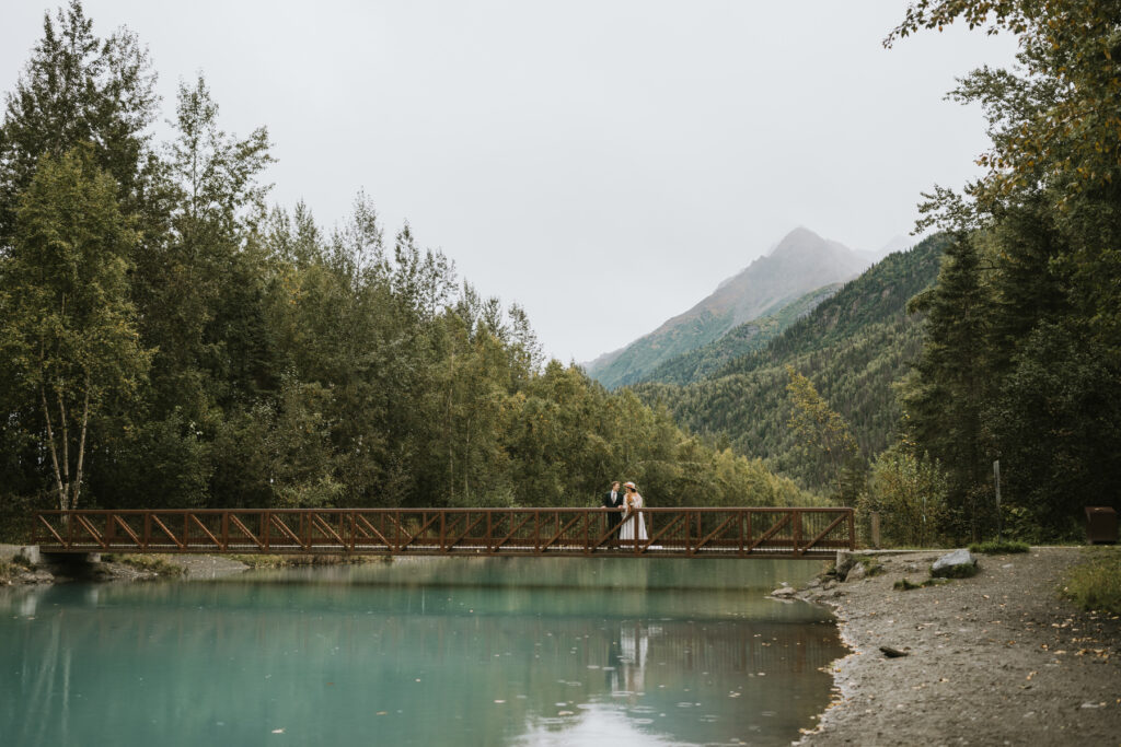 A couple stands on a wooden bridge over a turquoise river, surrounded by dense trees and mountains under a cloudy sky—a perfect scene for a lakeside elopement.