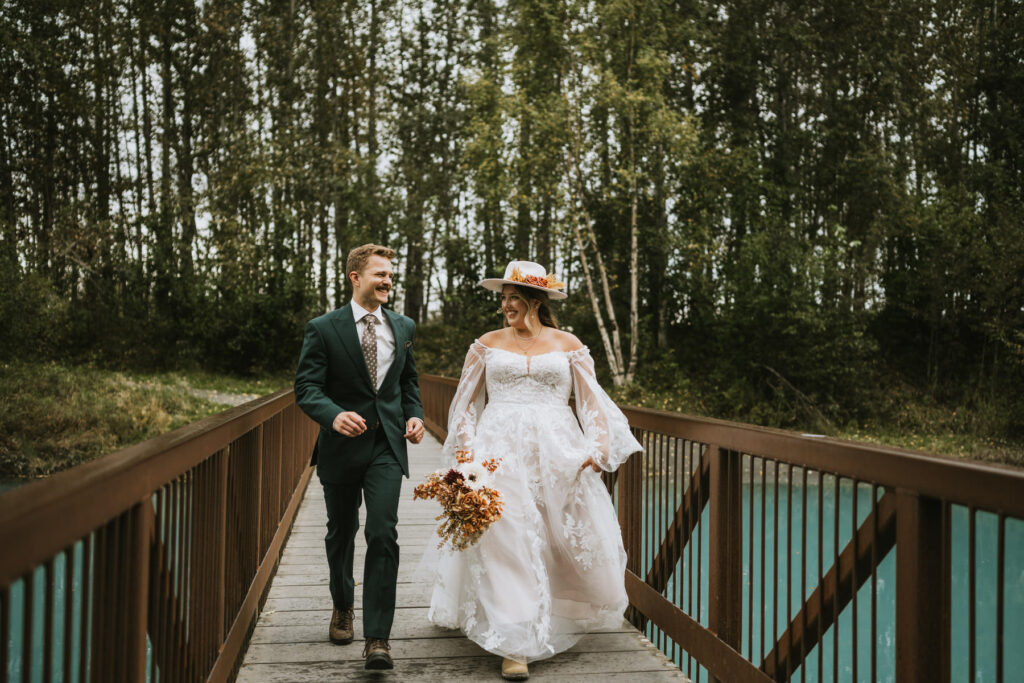 A bride and groom walk together on a wooden bridge over blue water in a lakeside elopement, surrounded by tall trees. The bride holds a bouquet and wears a white dress and hat; the groom wears a dark suit.
