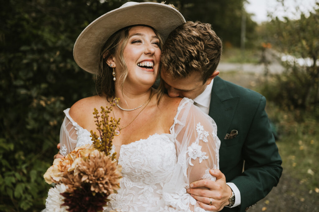 A bride in a white lace dress and hat holds a bouquet and laughs as the groom in a green suit embraces and kisses her shoulder during their lakeside elopement.