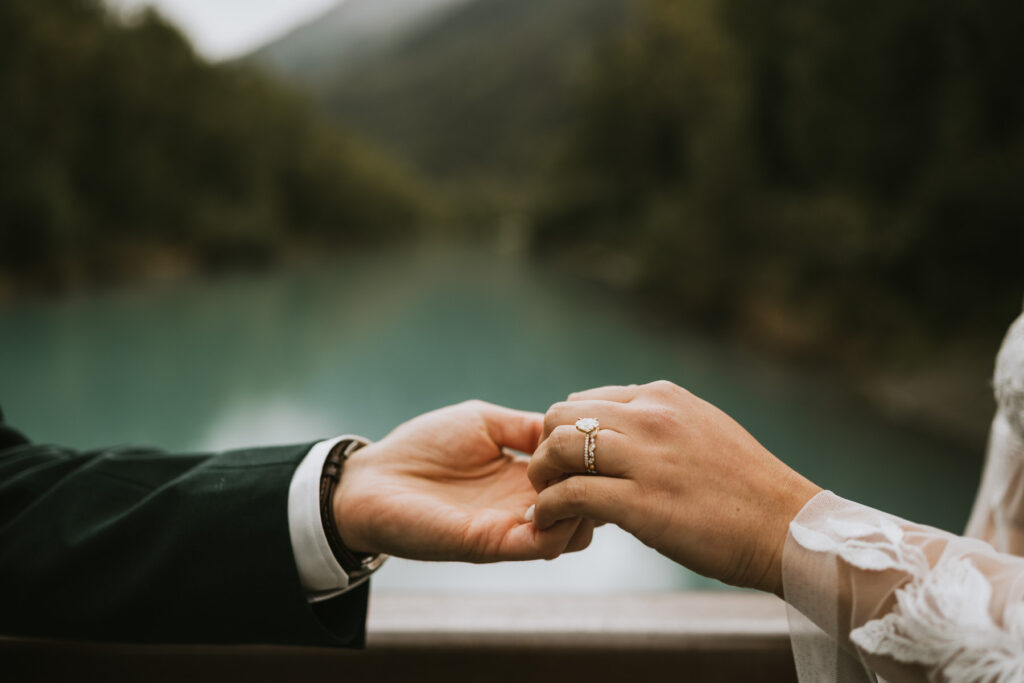 A person holds another's hand, showing an engagement ring during a lakeside elopement, with a blurred river and forested mountains in the background.