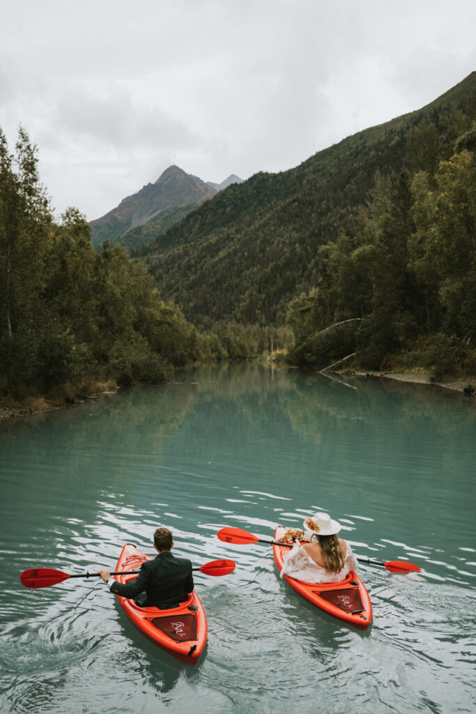 Two people paddle red kayaks on a calm, turquoise river surrounded by green forest and mountains under a cloudy sky, creating a perfect scene for a lakeside elopement.