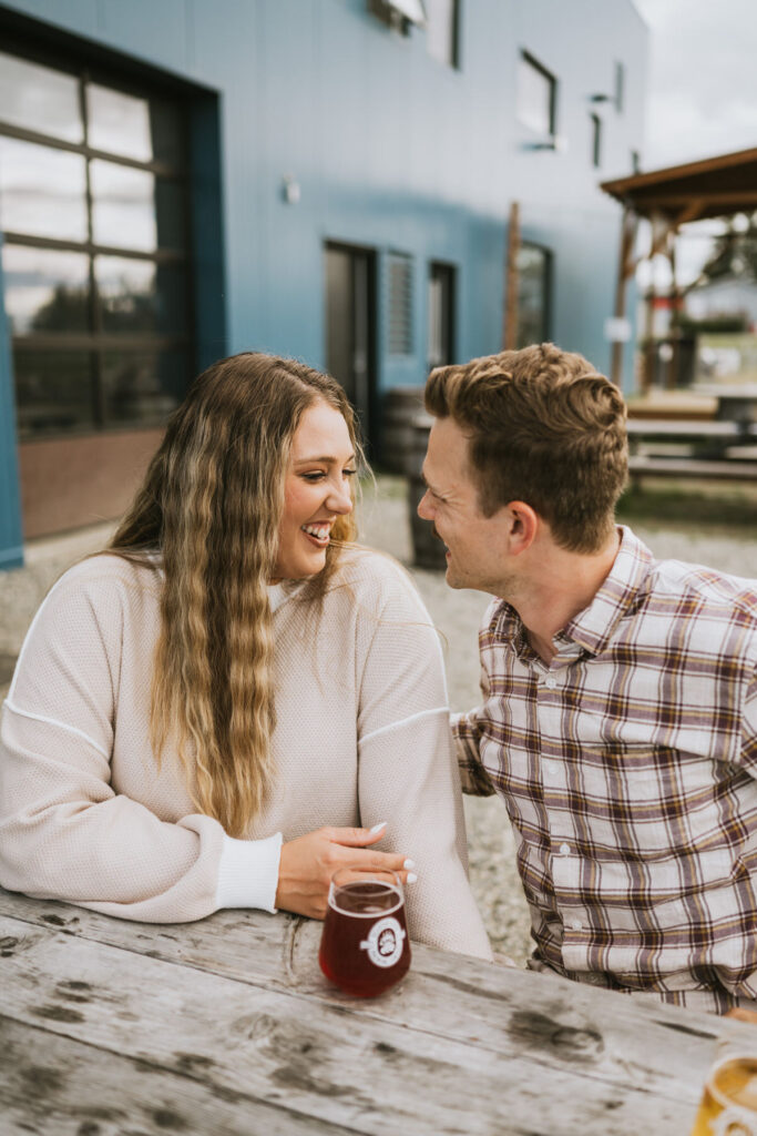 Two people sit at an outdoor wooden table, smiling at each other during their lakeside elopement. A glass of red beverage rests on the table, with a modern blue building in the background.