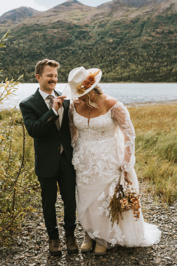 A bride in a lace dress and hat stands by the lake holding dried flowers during a lakeside elopement, while a groom in a suit playfully lifts the brim of her hat; mountains rise in the background.