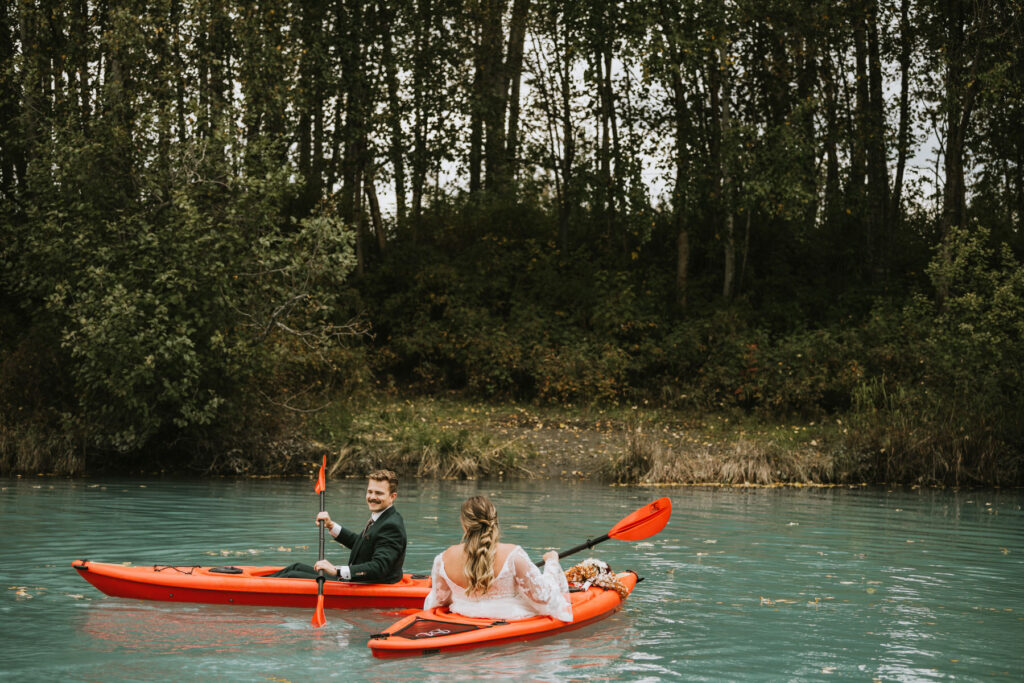 A couple in formal attire, including a white dress, enjoy a lakeside elopement as they paddle orange kayaks on a calm, turquoise river with a forested background.