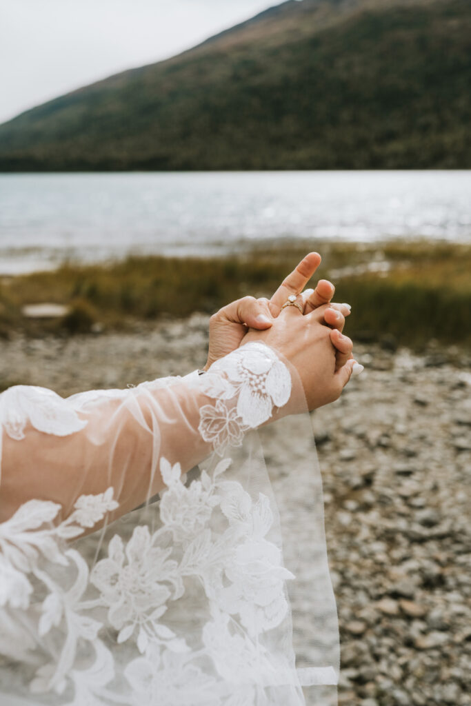 Close-up of a person wearing a white lace sleeve with floral patterns, hands clasped together near the lakeside—an intimate moment capturing the essence of a lakeside elopement with mountains in the background.