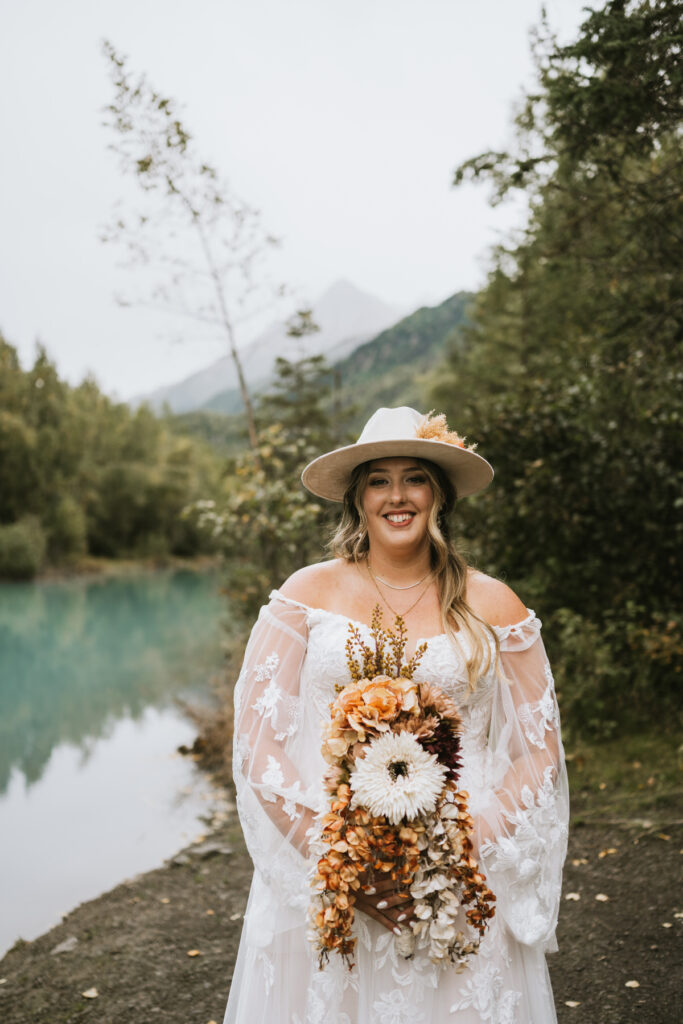 A woman in a white lace dress and wide-brimmed hat holds a cascading bouquet of flowers, standing outdoors by the water—a dreamy lakeside elopement with trees and mountains as a stunning backdrop.