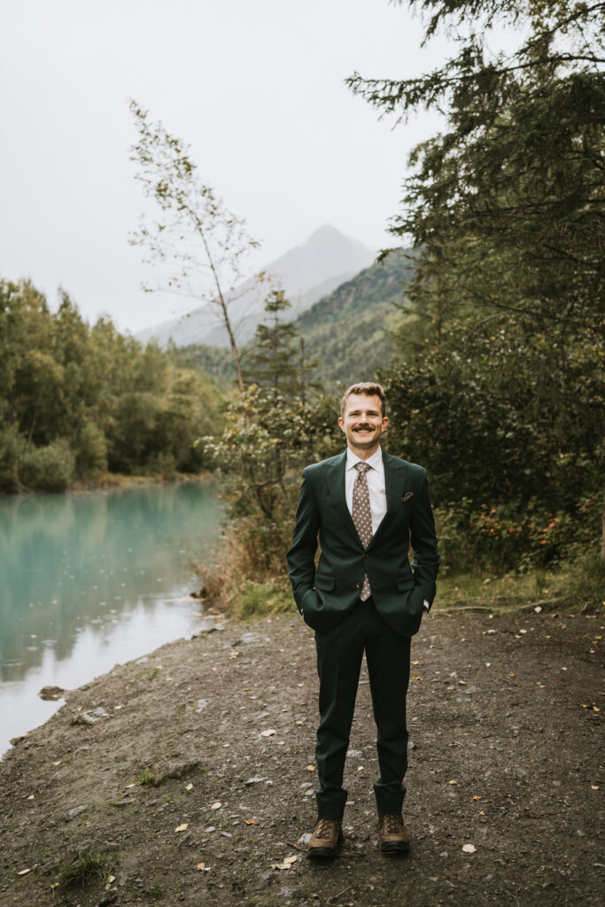 A man in a dark suit and tie stands on a dirt path beside a turquoise lake, setting the scene for an intimate lakeside elopement, with trees and a mountain in the background.