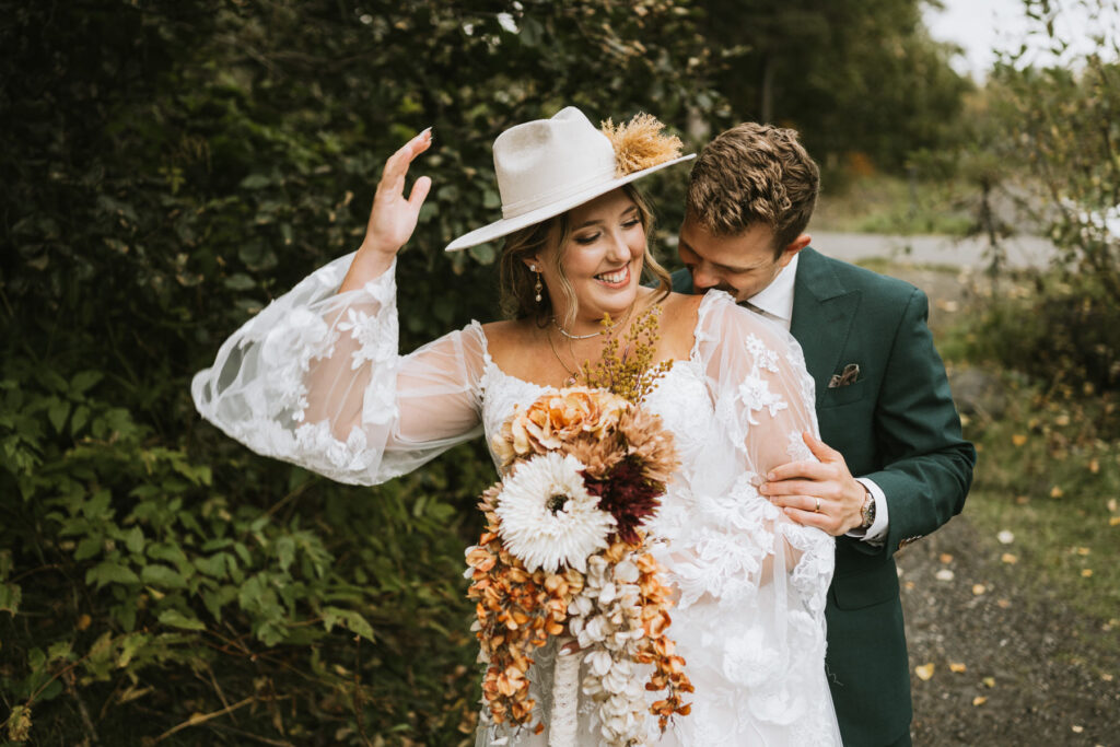 A bride in a white lace dress and hat holds a bouquet as a groom in a green suit embraces her from behind outdoors during their lakeside elopement, both smiling.