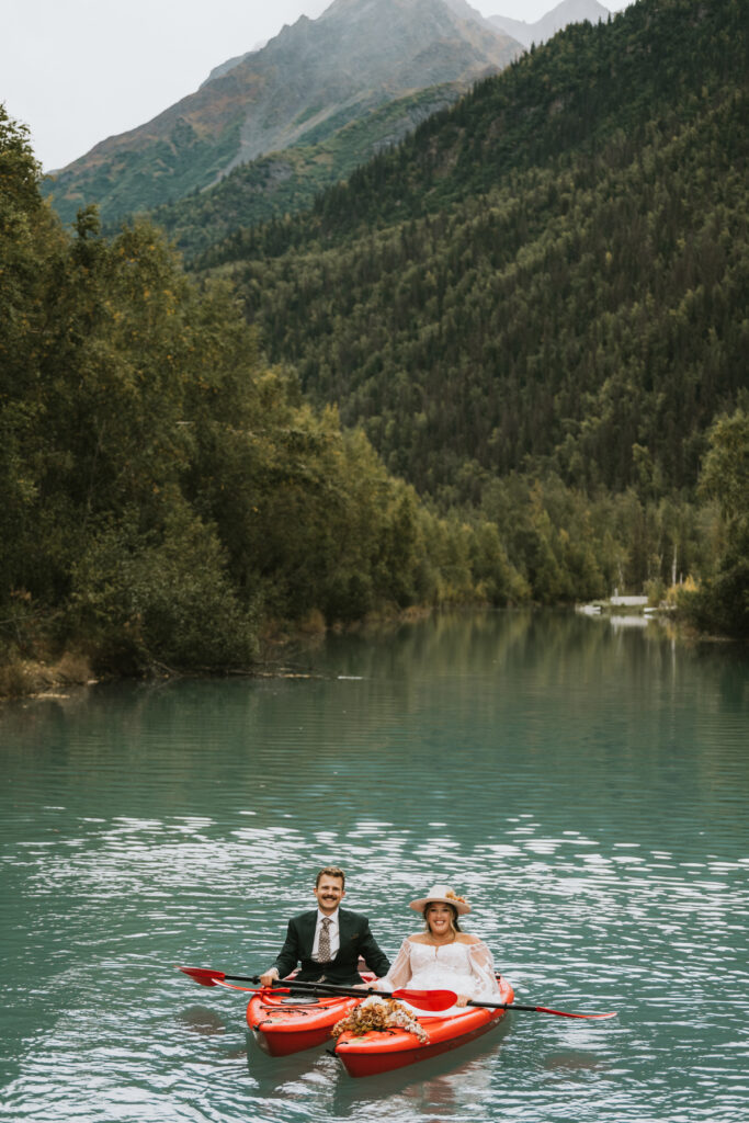 A couple dressed in wedding attire sits in red kayaks on a calm river, enjoying a lakeside elopement surrounded by green trees and mountains in the background.