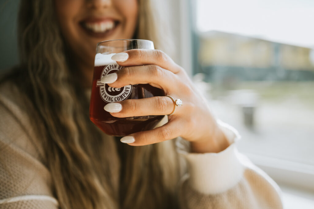 A person with long nails and a ring on their finger holds a glass of beer, smiling inside a bright room, reminiscing about their joyful lakeside elopement.