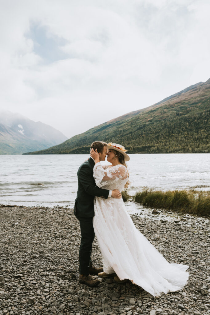 A bride and groom embrace and kiss on a rocky lakeshore, celebrating their lakeside elopement with mountains and trees in the background.