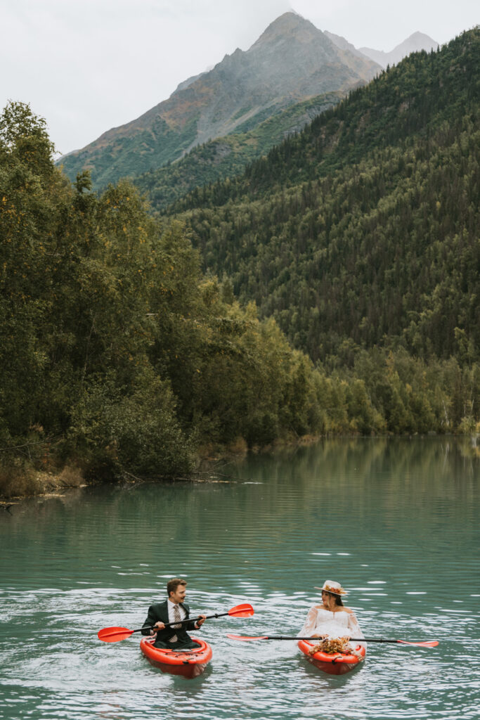 Two people in wedding attire paddle red kayaks on a calm lake, surrounded by dense trees and mountains under a cloudy sky—a picture-perfect lakeside elopement.