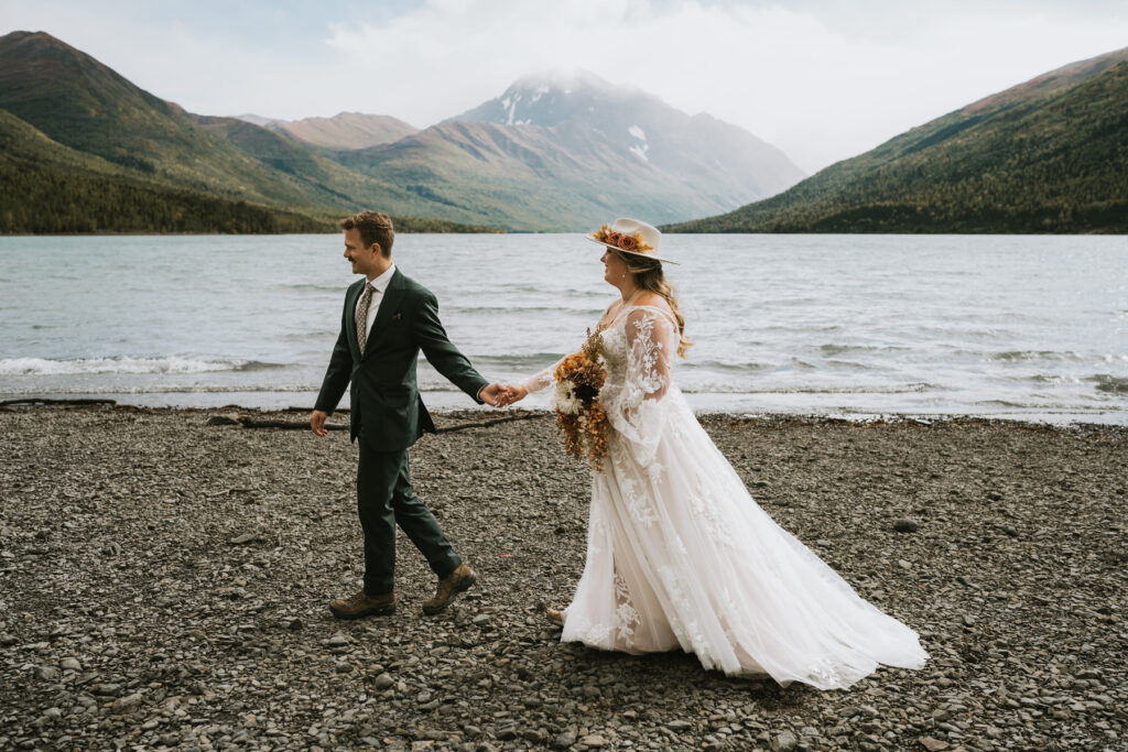 A couple in wedding attire walks hand in hand along a rocky lakeshore, capturing the magic of a lakeside elopement with mountains and shimmering water in the background.