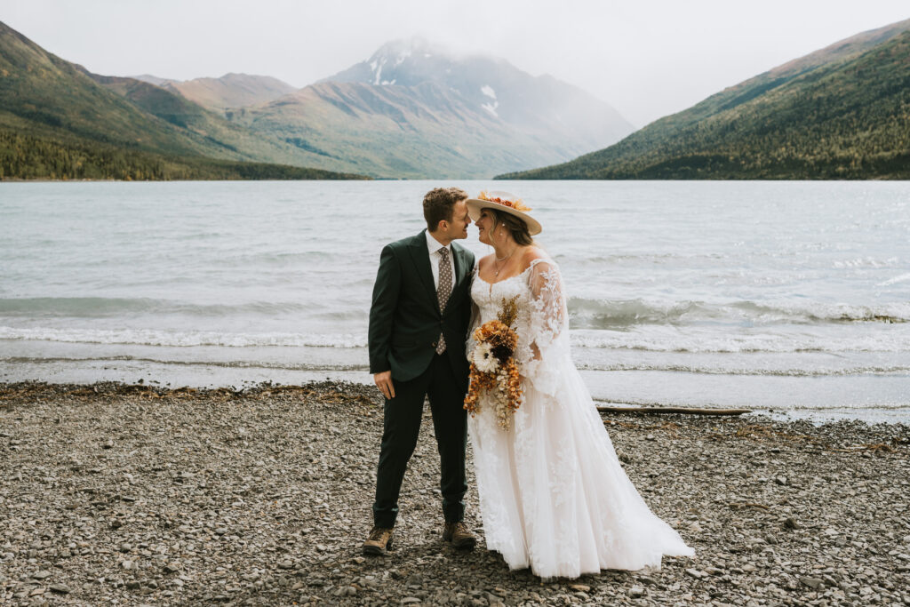 A bride and groom stand together on a rocky lakeshore, embracing during their intimate lakeside elopement with majestic mountains and sparkling water in the background.