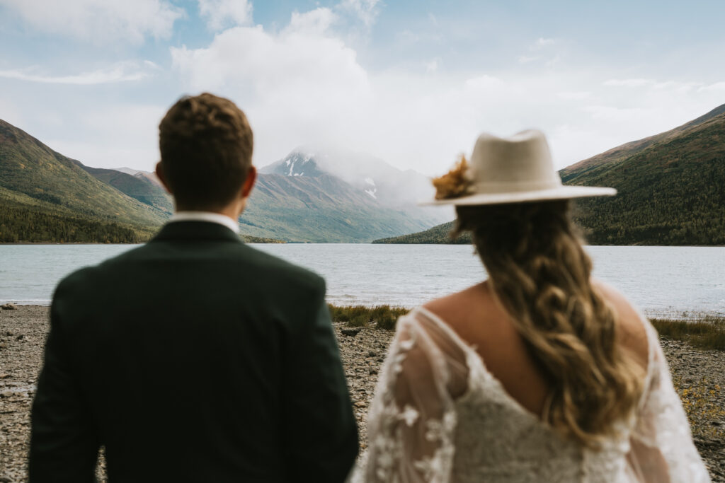 A couple in wedding attire stands on a rocky shore during their lakeside elopement, facing a tranquil lake with mountains and clouds in the background.