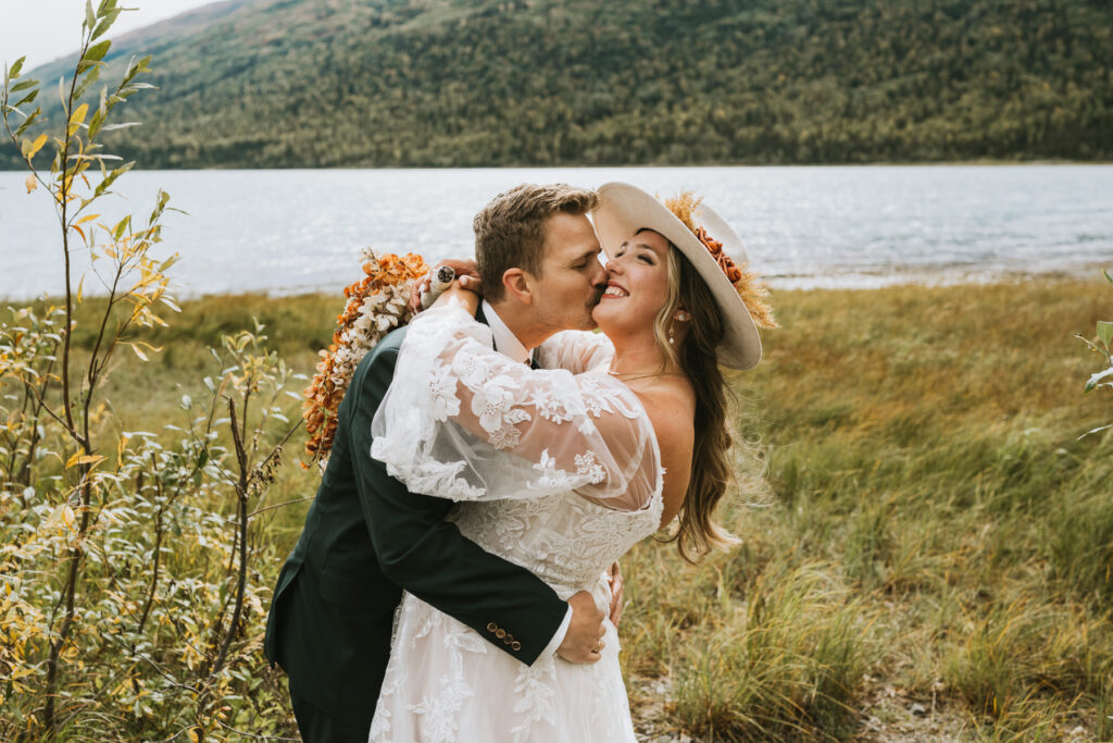 A bride in a white dress and hat smiles as the groom, in a dark suit, kisses her cheek during their lakeside elopement near scenic mountains.
