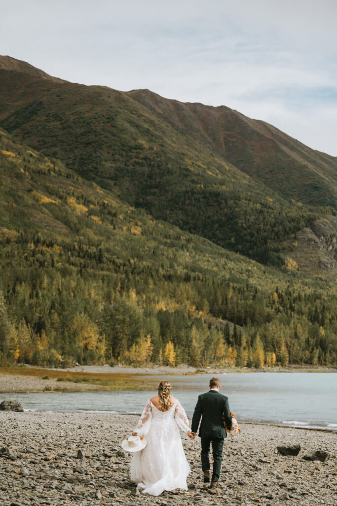 A bride and groom walk hand in hand along a rocky lakeshore, forested mountains rising behind them, savoring a romantic lakeside elopement.