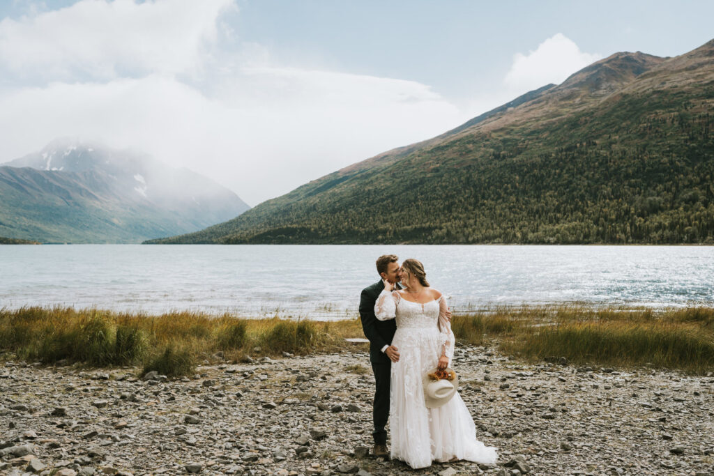 A bride and groom stand on a rocky lakeshore, surrounded by mountains and water, celebrating their lakeside elopement as the bride holds a hat and bouquet.