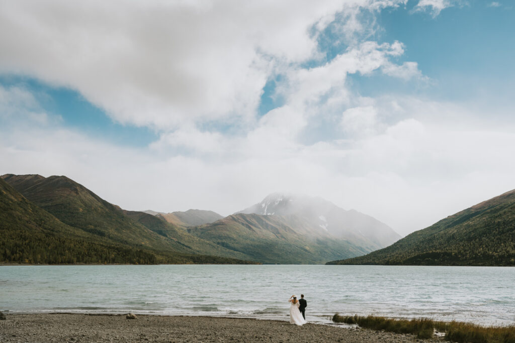 A couple in wedding attire stands on the shore of a lake, surrounded by mountains under a partly cloudy sky, capturing the magic of a lakeside elopement.
