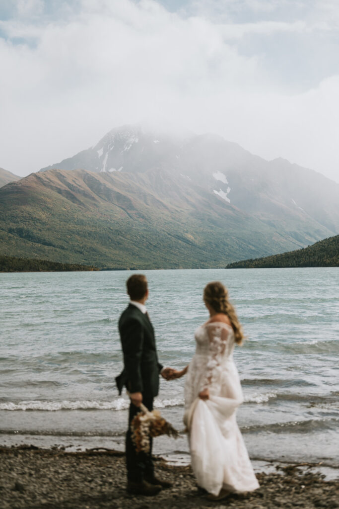 A bride and groom stand hand in hand on a rocky shore during their lakeside elopement, facing a tranquil lake with mountains rising in the background beneath a cloudy sky.