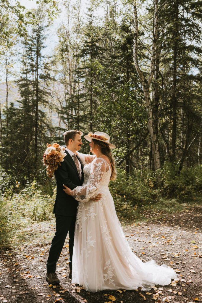 A bride and groom stand together in a forest for their lakeside elopement, facing each other as the bride holds a bouquet and wears a lace wedding dress and hat.