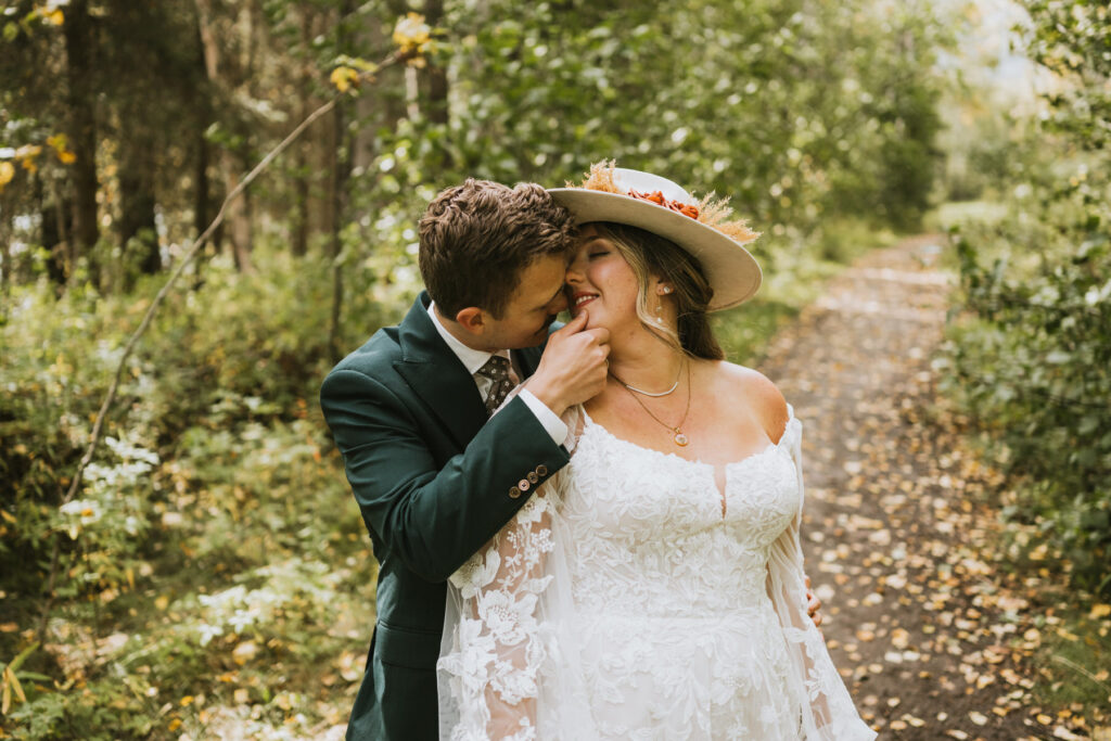 A bride in a white lace dress and hat stands on a wooded path during their lakeside elopement, while the groom in a green suit kisses her cheek and holds her hand to his lips.