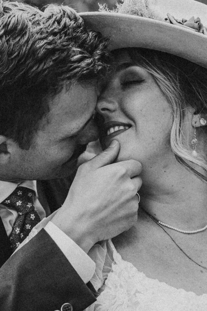A close-up, black and white photo of a couple embracing at their lakeside elopement; the man gently touches the woman's chin, both smiling with closed eyes.