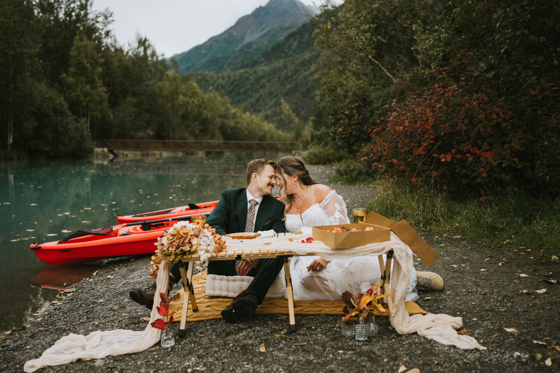 A couple dressed in wedding attire enjoys a lakeside elopement, sitting on a picnic blanket by the lake with red kayaks, a table of food, and mountains in the background.