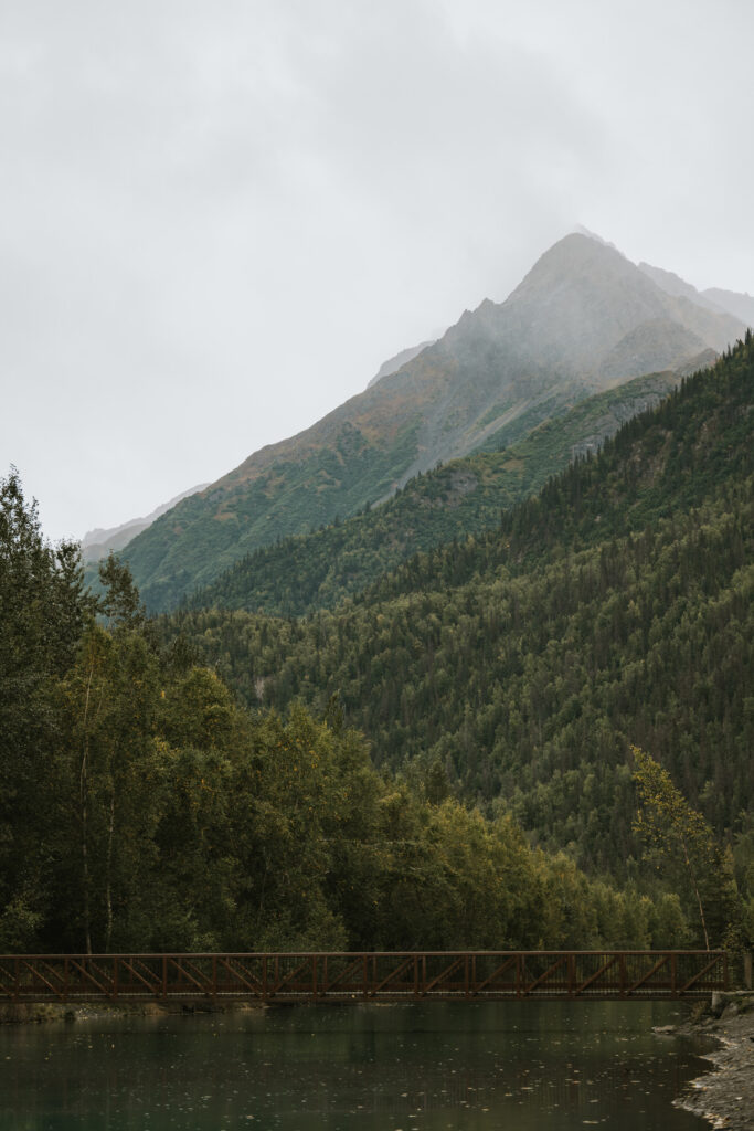A wooden bridge spans across a river in the foreground, perfect for a lakeside elopement, with a densely forested mountain rising in the background under a cloudy sky.