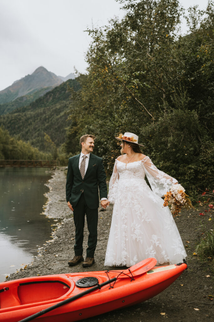 A bride and groom stand by a lake holding hands, a red kayak on the shore beside them, mountains rising behind—capturing the magic of their lakeside elopement.