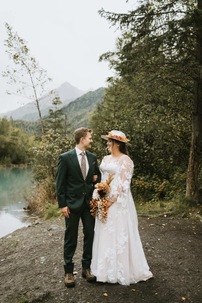 A bride and groom stand outdoors by a lake and trees, facing each other during their lakeside elopement. The bride holds a bouquet and wears a floral hat; the groom is in a dark suit and boots.