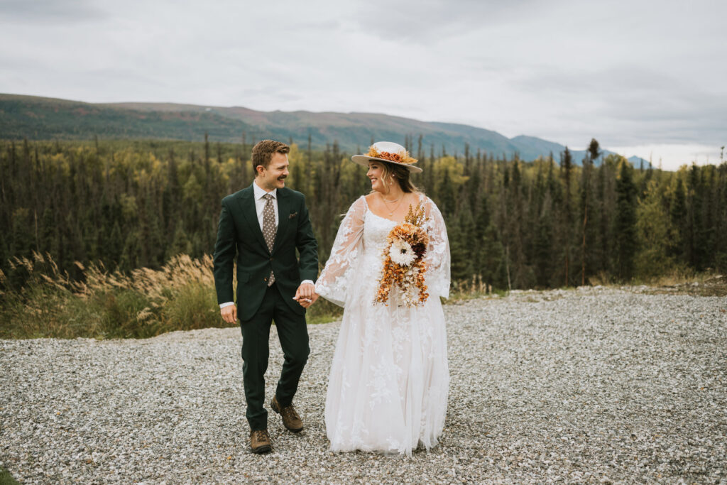 A bride and groom in wedding attire walk hand-in-hand outside on a gravel path, with trees, mountains, and the tranquil beauty of a lakeside elopement in the background.