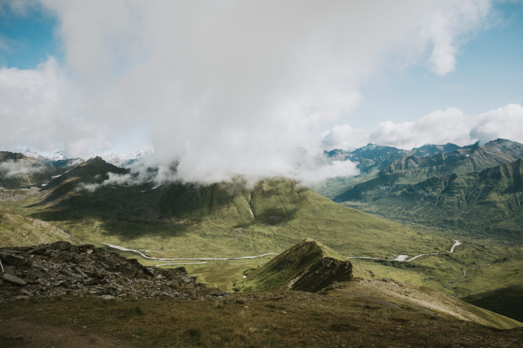 A green mountainous landscape with a winding road and river, partly covered by low clouds under a partly cloudy sky—a scene reminiscent of breathtaking Alaska hikes.