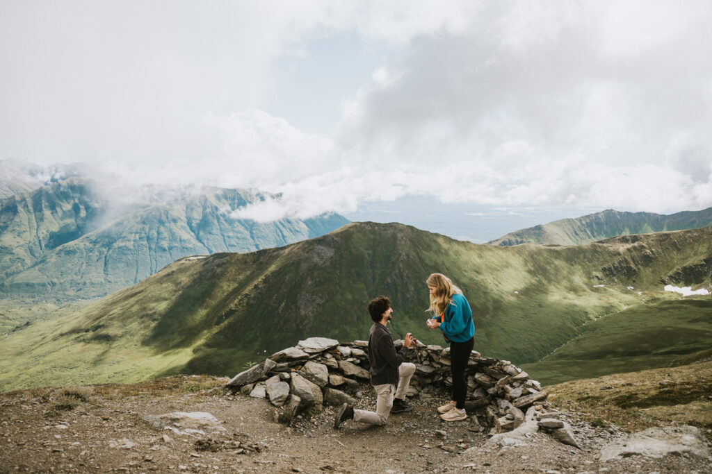 A person kneels and proposes to another on a mountain viewpoint along one of the scenic Alaska hikes, surrounded by stone, with green hills and clouds in the background.