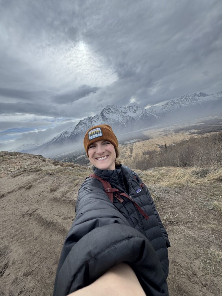 Person smiling and taking a selfie on a dirt trail during one of their favorite Alaska hikes, with snow-capped mountains and a cloudy sky in the background.