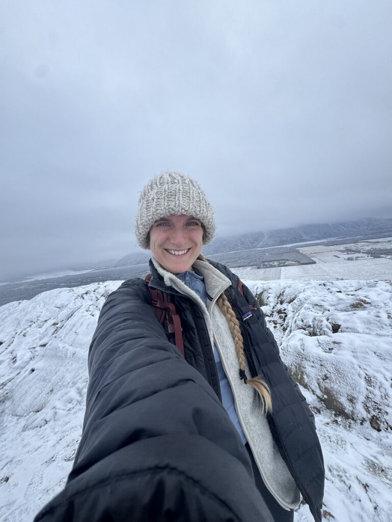 A person wearing a knit hat and winter coat smiles at the camera while standing on a snow-covered landscape under a cloudy sky, ready to explore scenic Alaska hikes.