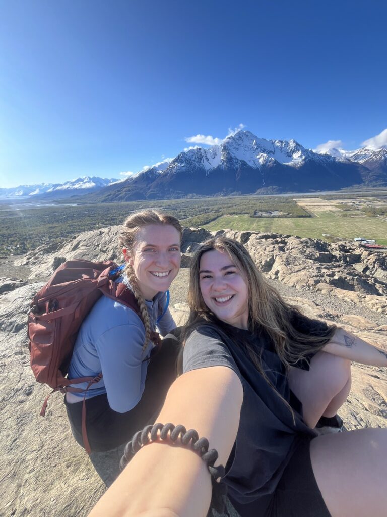 Two people sit on a rocky ledge, smiling for a selfie with snow-capped mountains and a clear blue sky in the background—a perfect moment from one of their unforgettable Alaska hikes.
