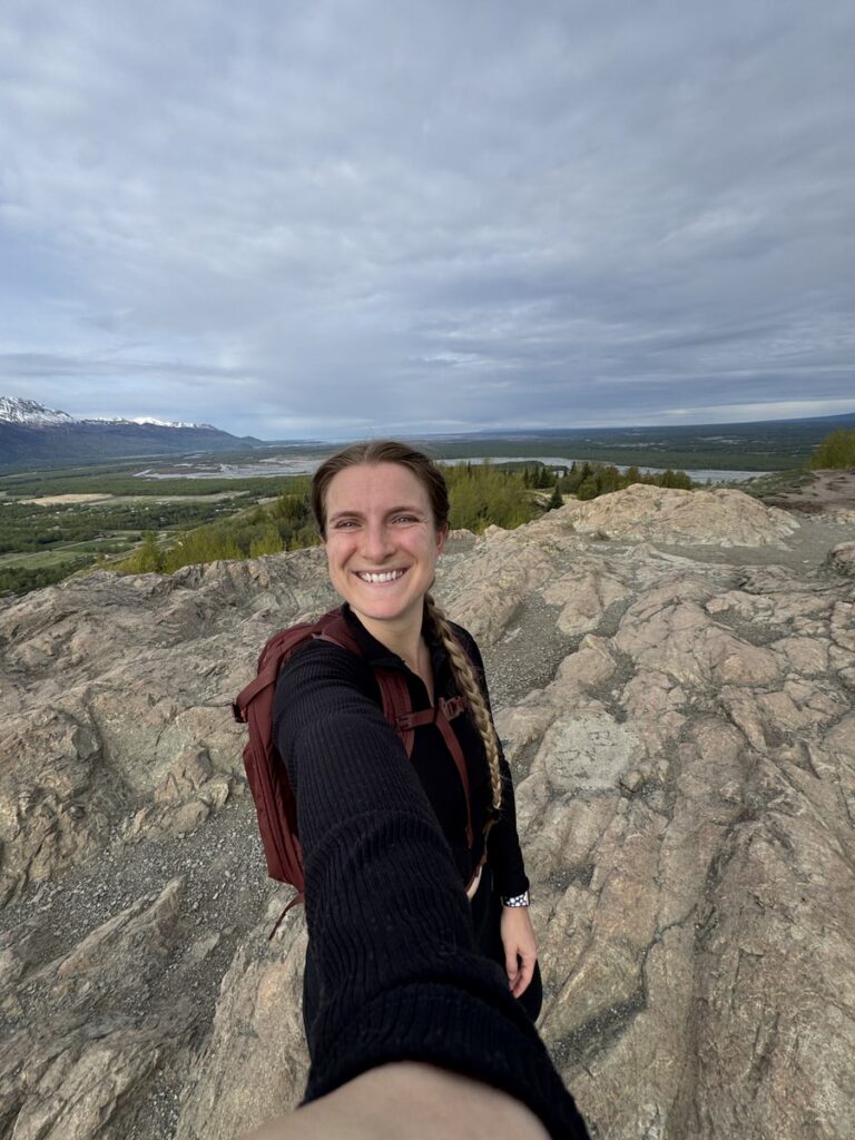 A person with a backpack smiles while taking a selfie on a rocky hilltop, capturing the spirit of Alaska hikes with a vast landscape of trees and distant mountains in the background under a cloudy sky.